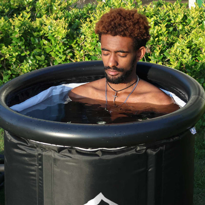 Relaxing man in a matte black portable cold plunge tub outdoors amidst green foliage. Ideal for cold plunge recovery.