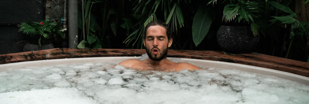 Man enjoying cold plunge therapy session in white ice bath tub surrounded by tropical plants.