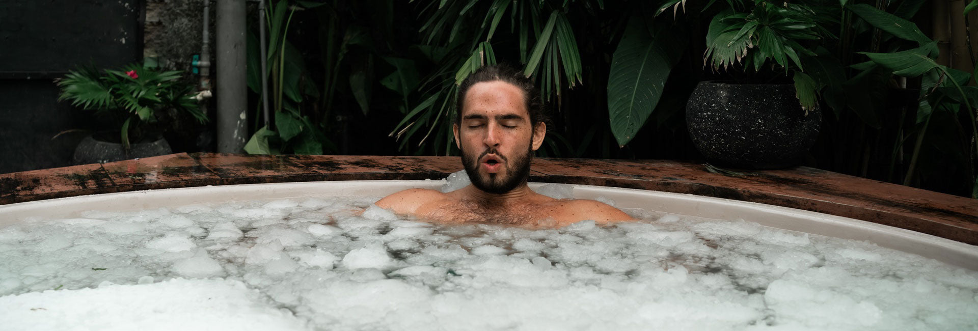Man enjoying cold plunge therapy session in white ice bath tub surrounded by tropical plants.