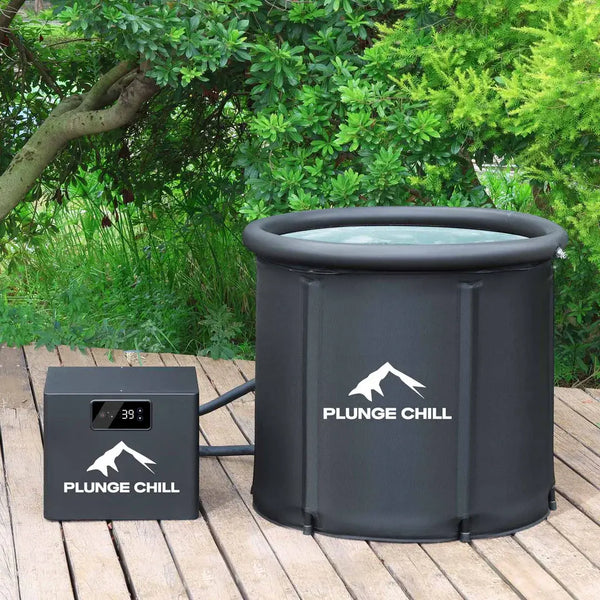 A large, cylindrical, black Plunge Chill ice bath tub with an inflatable upper ring sits on a wooden deck outdoors, surrounded by lush green foliage. To the left is a smaller, matching black control unit/chiller, also branded Plunge Chill, with a digital temperature display showing 39°F.. The entire setup is ready for cold water immersion.