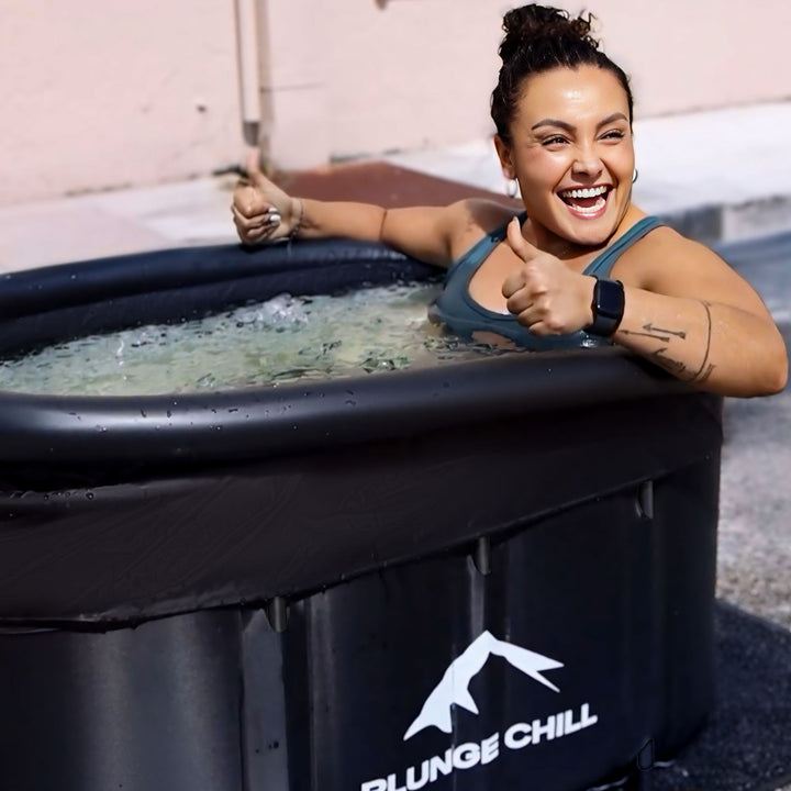 A smiling woman giving thumbs up while sitting in a black Plunge Chill cold plunge tub outdoors.