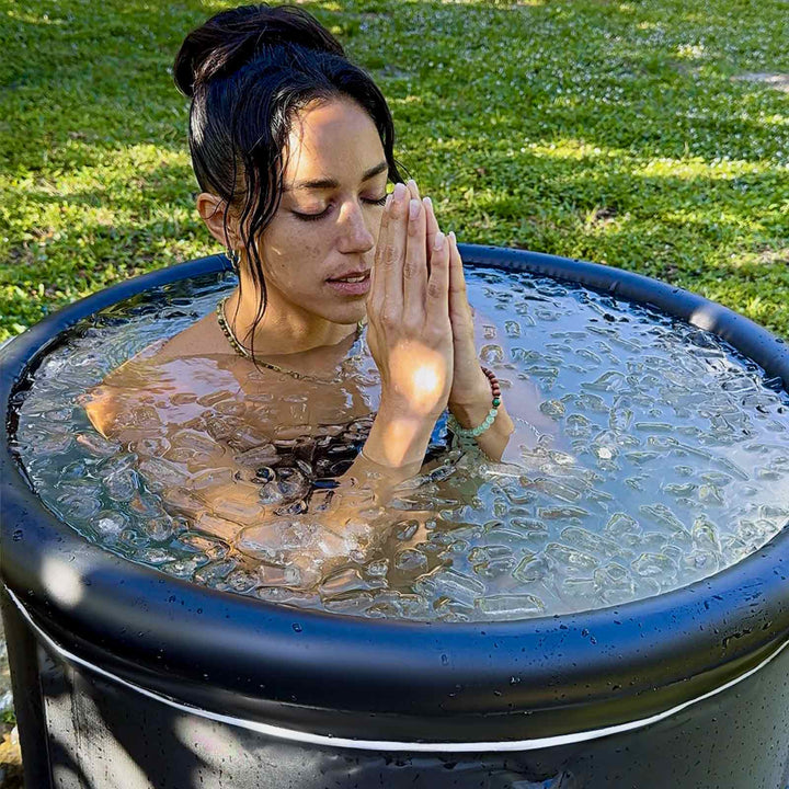 A Female sit in a black portable & inflatable cold plunge tub outdoors amidst green foliage. Ideal for cold plunge recovery.